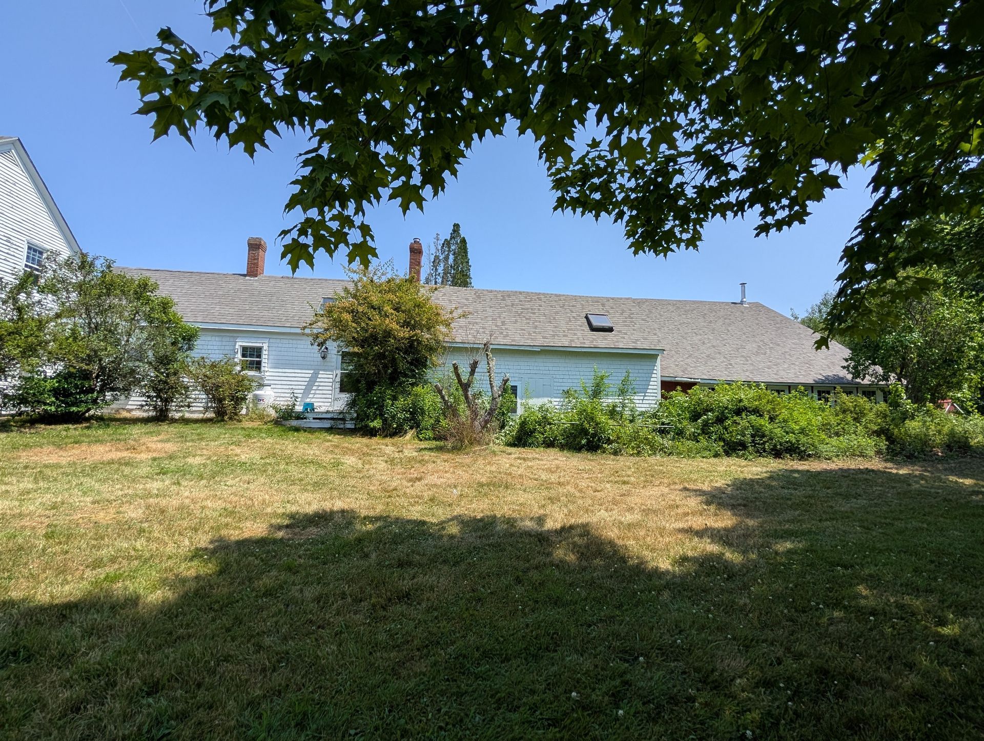A long, light-blue house with a grey shingled roof stands behind a grassy yard on a sunny day.