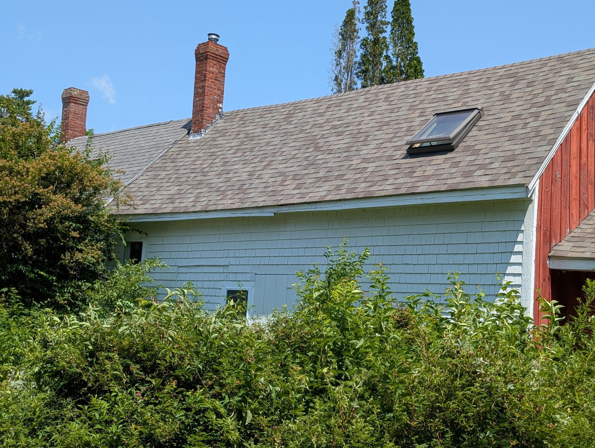 A white-sided house with two brick chimneys and a skylight on its gray-shingled roof, partially obscured by green bushes.