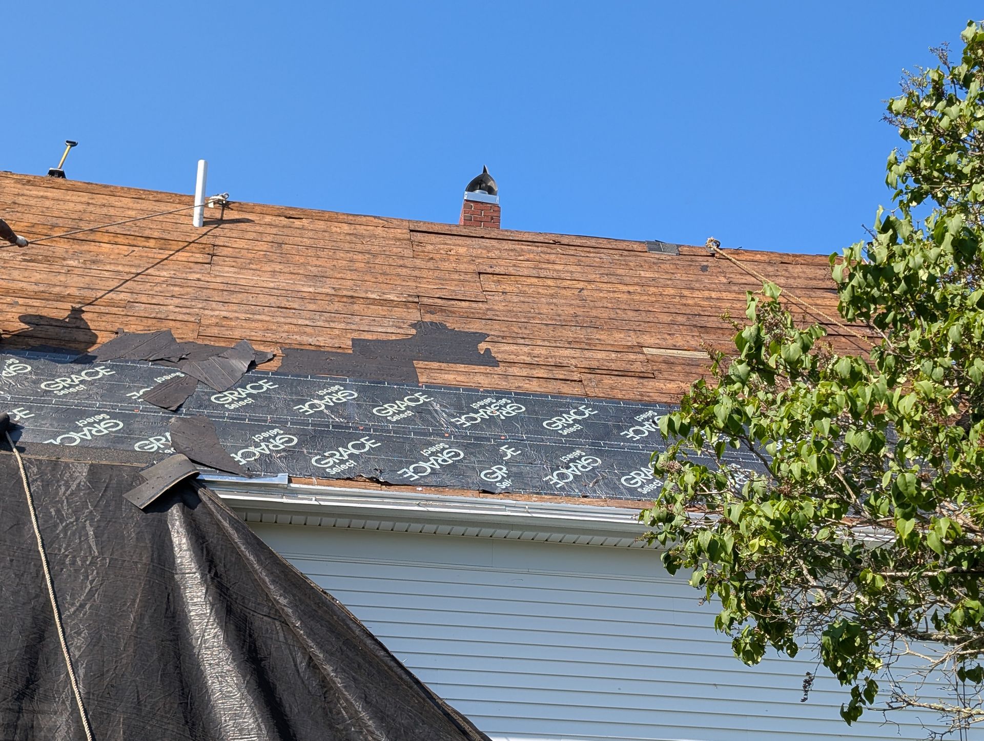 A residential roof under repair with sections of shingles removed, exposing the black underlayment and a brick chimney.