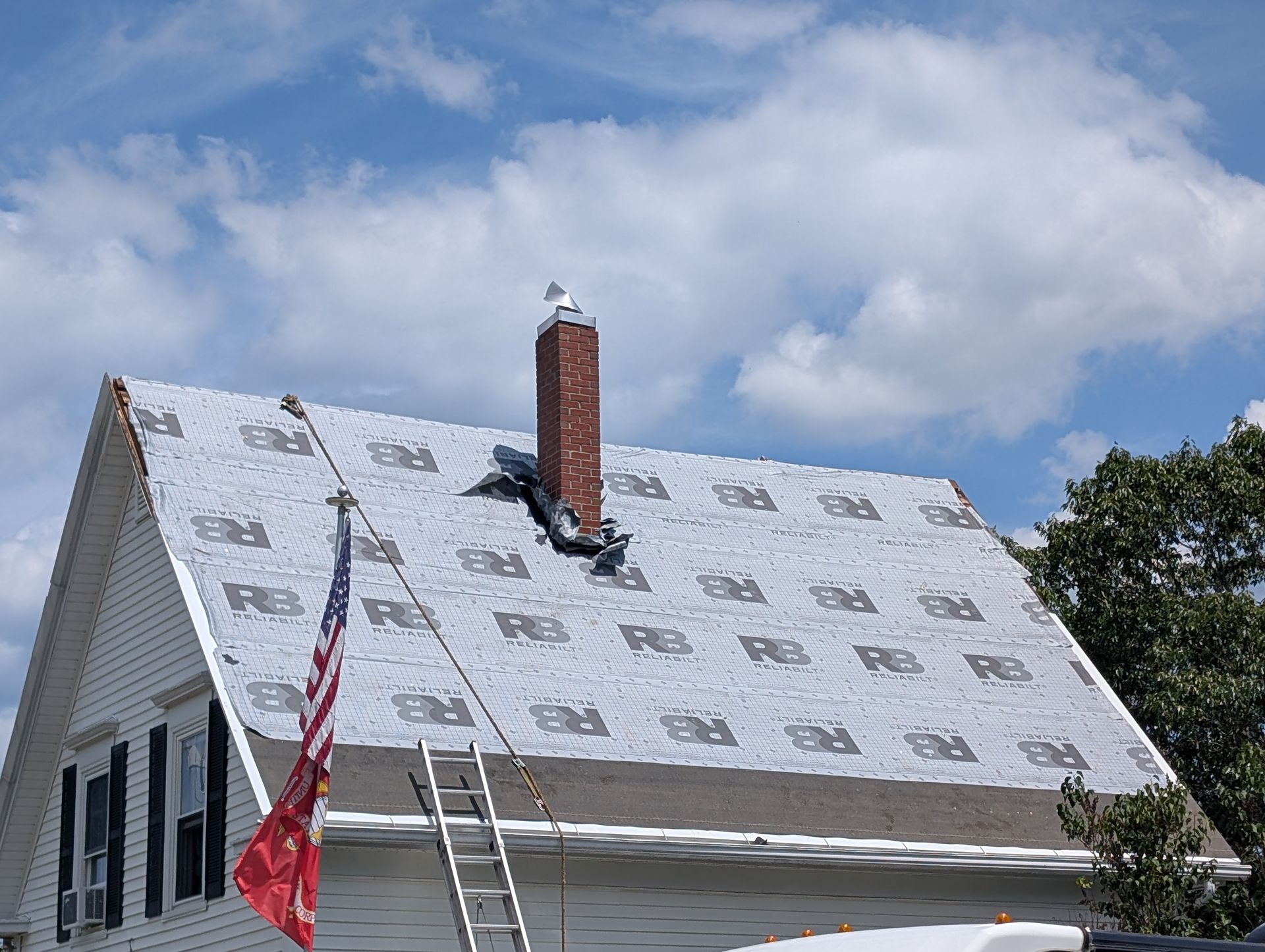 A roof undergoing repairs, covered in gray underlayment with a brick chimney and an American flag attached to a ladder.