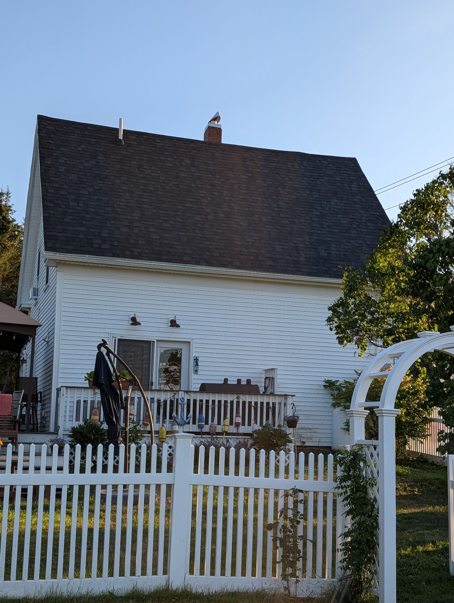 A white house with a dark gabled roof and a wooden fence, seen from a garden with an arched trellis on a sunny day.