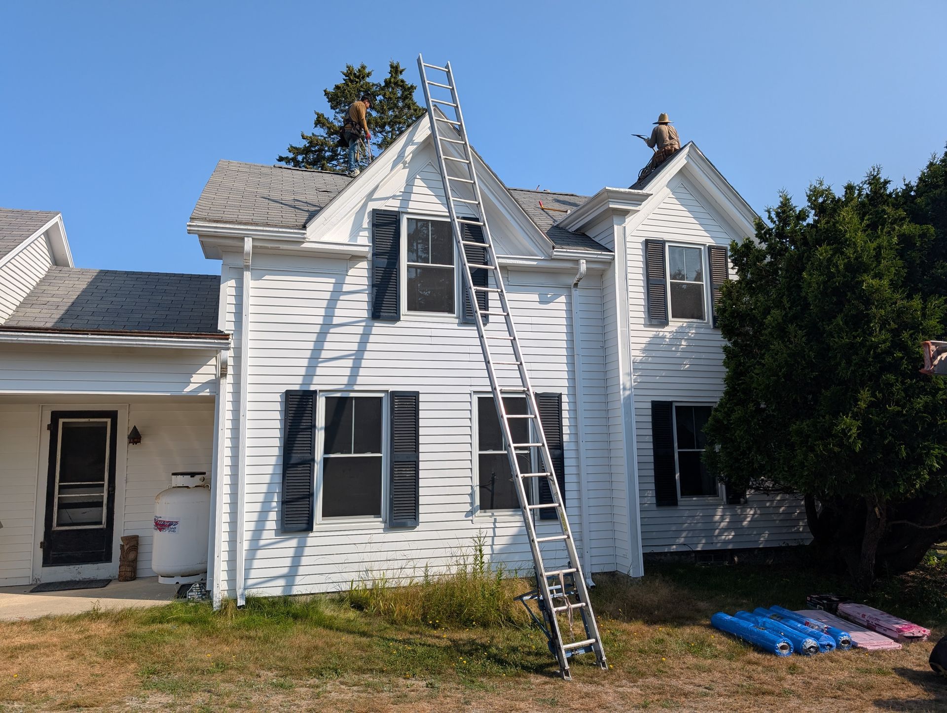 A two-story white house with two workers on the roof and a tall aluminum extension ladder leaning against the front.