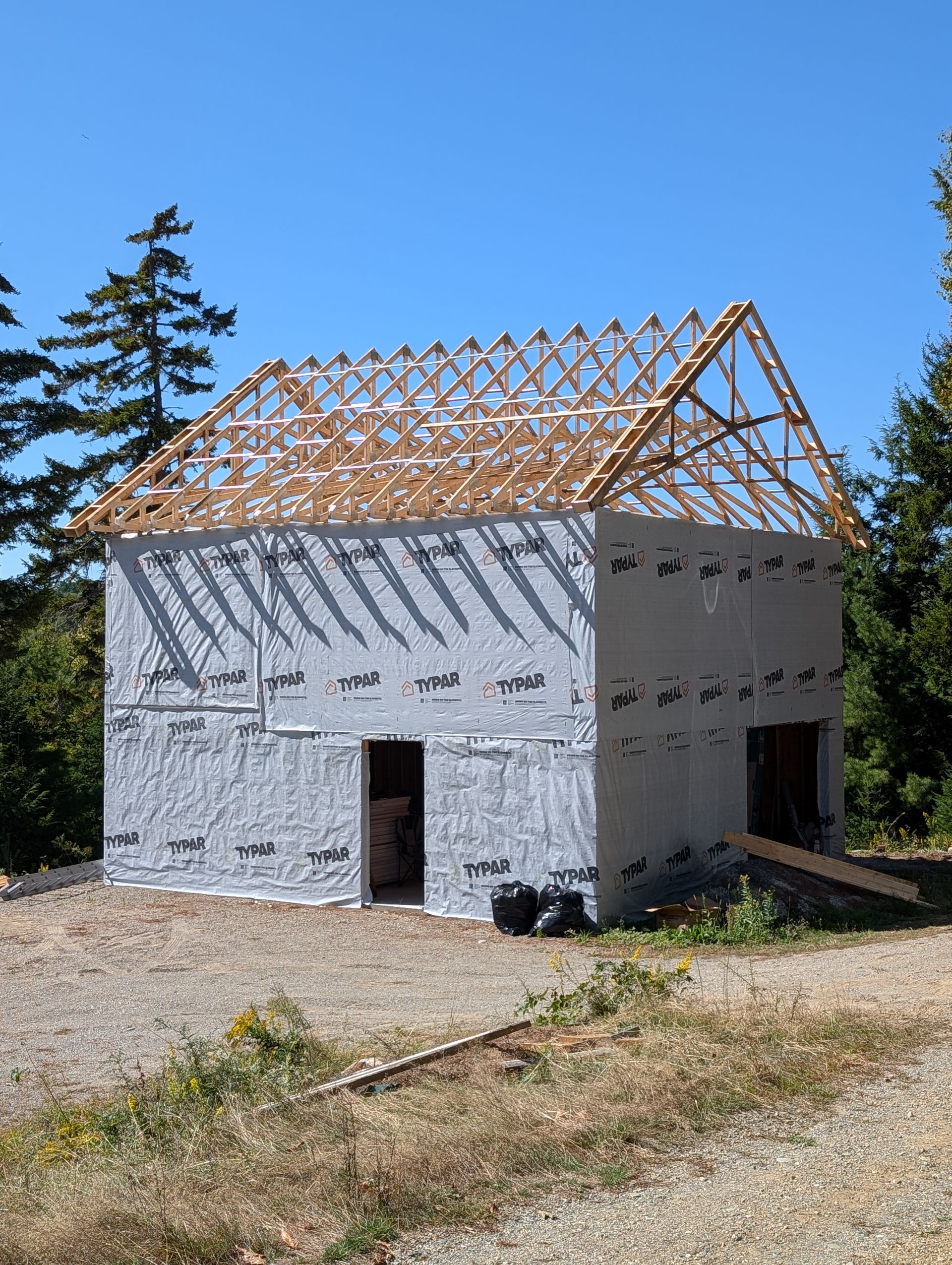 A partially constructed two-story building featuring an exposed wooden roof truss frame, exterior sheathing, and trees.