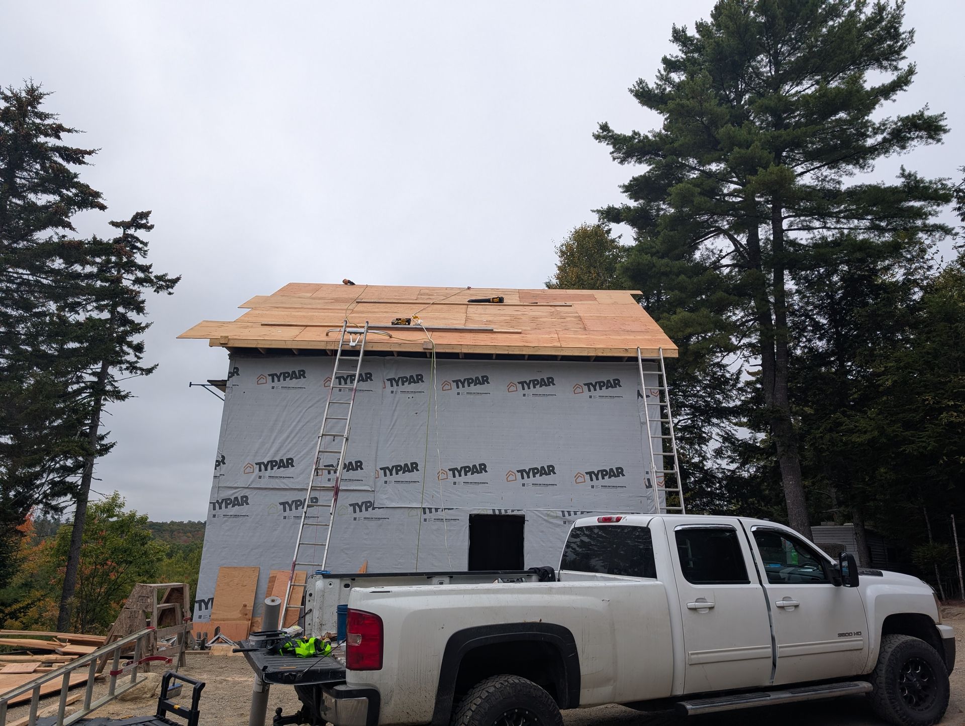 A white pickup truck parked in front of a house under construction with exposed roof sheathing and gray house wrap.