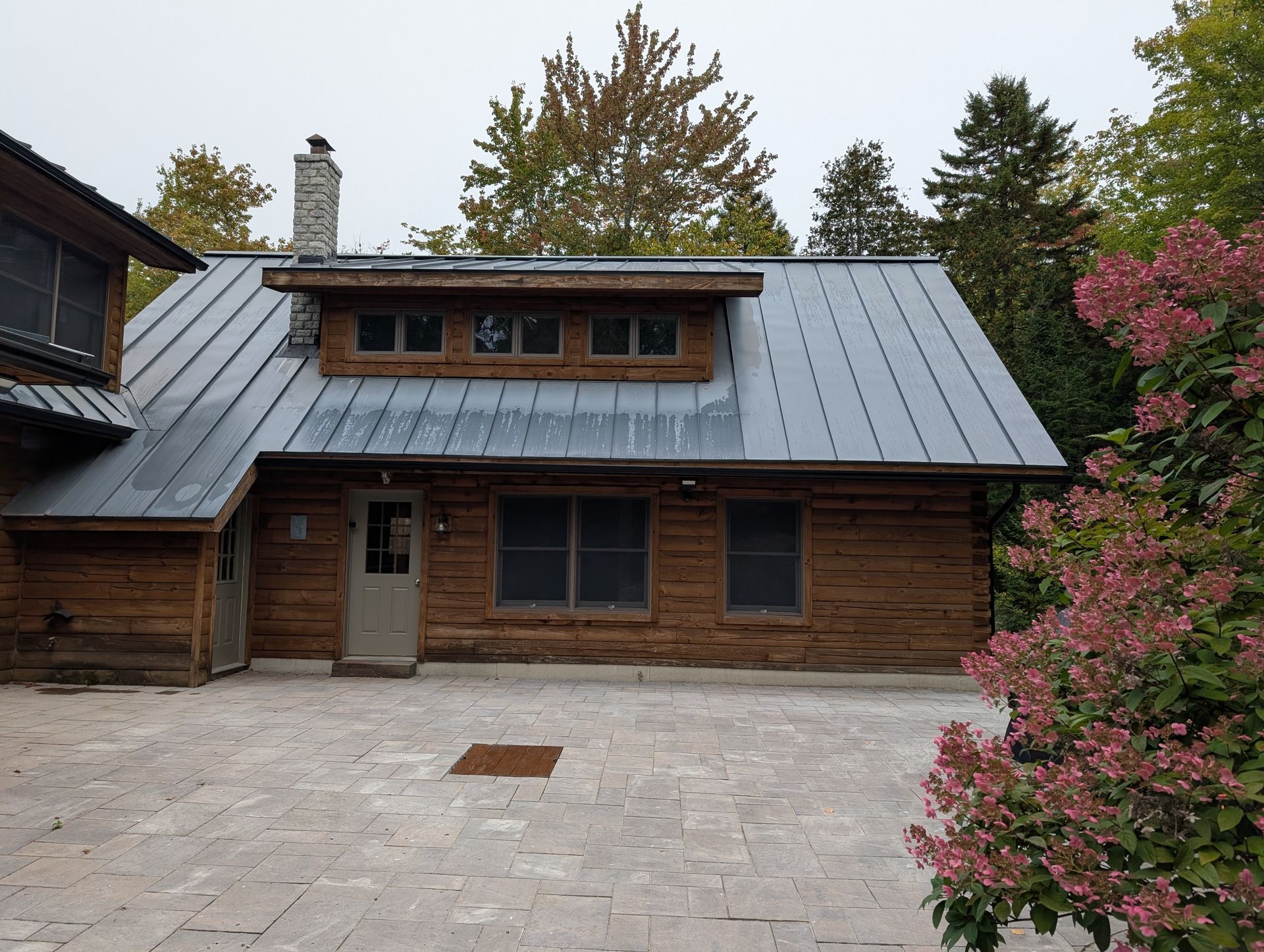 A rustic log cabin with a gray metal roof, a stone chimney, and a stone patio, surrounded by trees and a flowering bush.