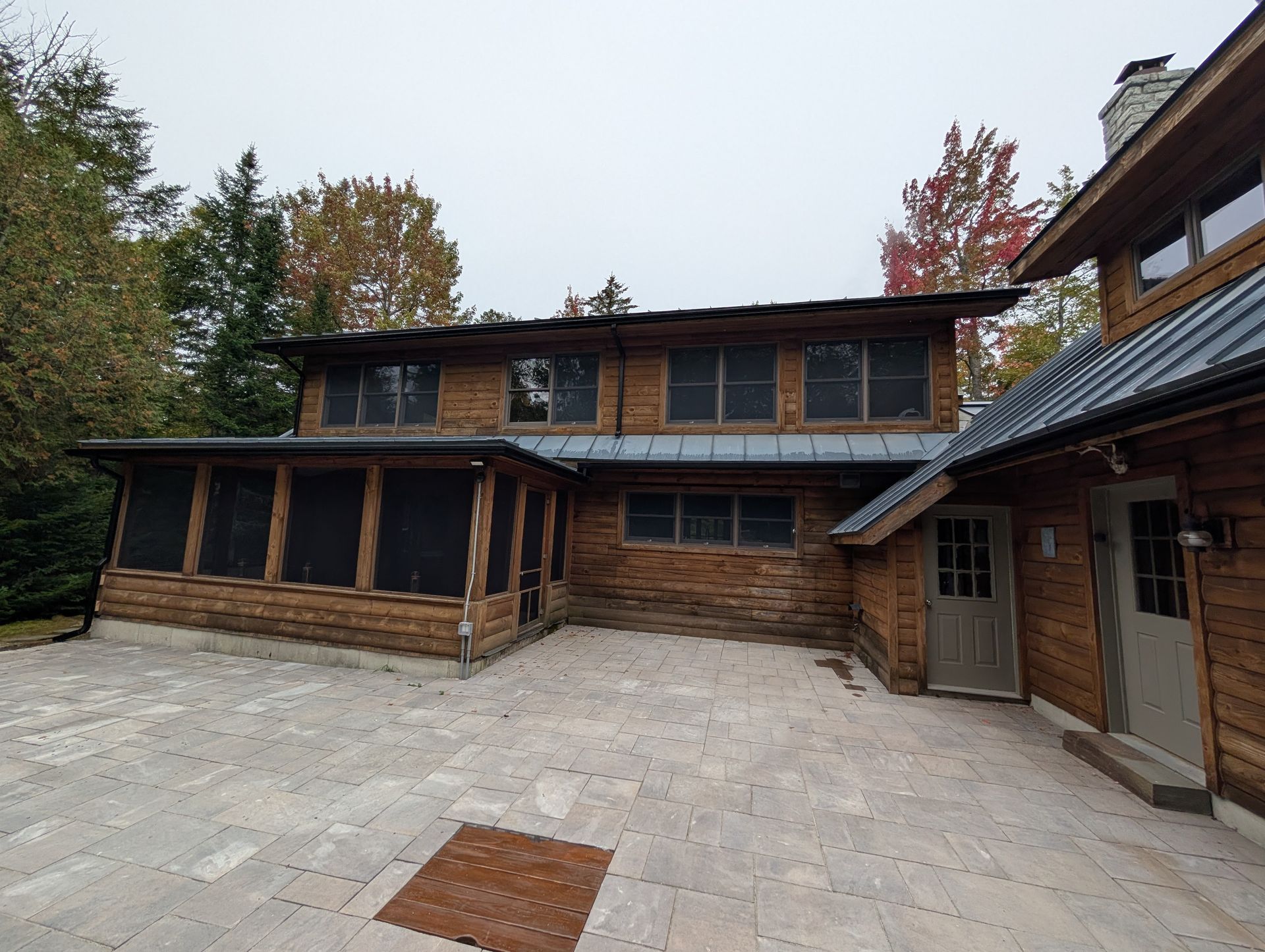 A stone patio leads to a two-story wooden cabin with a screened-in porch and grey metal roofing under an overcast sky.