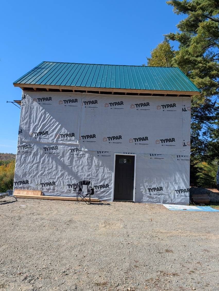 A two-story building under construction, wrapped in gray Tyvek house wrap with a green metal roof under a blue sky.