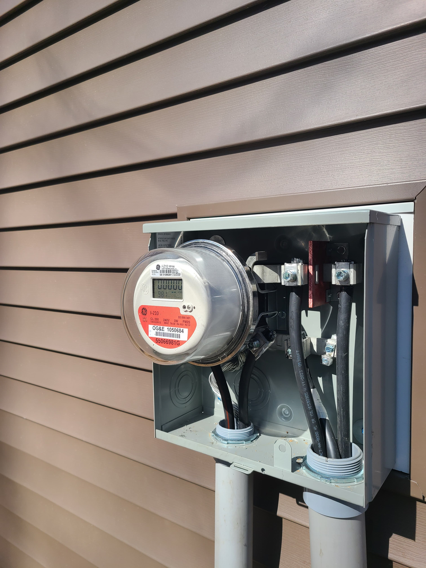 Electric meter in an open gray utility box mounted on tan house siding.