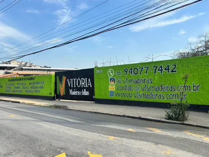 Placa verde e preta com o nome do edifício Vitória Madeiras, com informações de contacto, sob um céu azul.