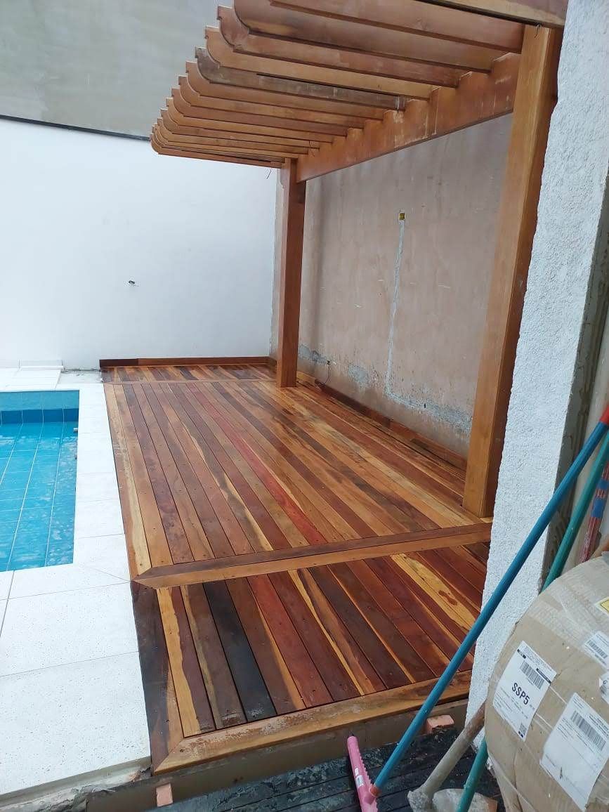Deck de madeira com pérgola ao lado da piscina. Madeira marrom, parede clara, água azul.