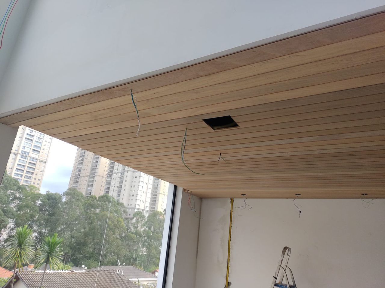 Teto de madeira com uma abertura quadrada, rachaduras e fios, acima de uma janela com vista para a cidade.