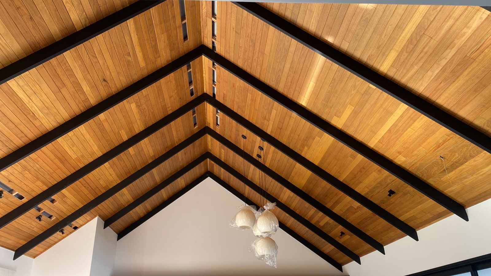 Teto de tábuas de madeira com vigas pretas em uma sala branca com teto abobadado.