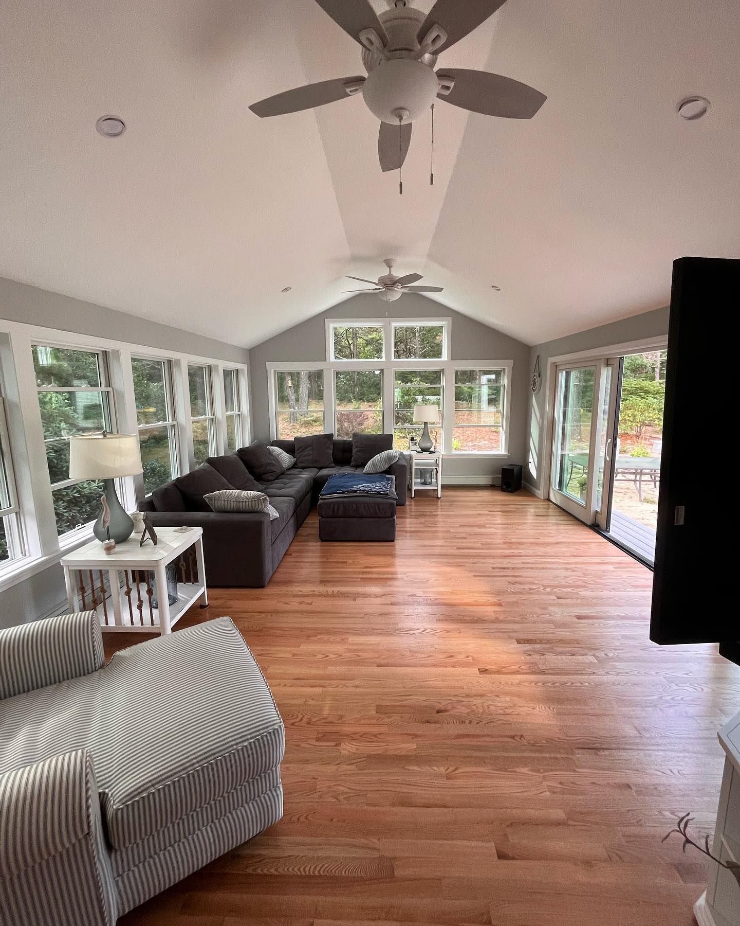 Bright sunroom with hardwood floors, gray sectional sofa, chairs, ceiling fans, and large windows overlooking greenery