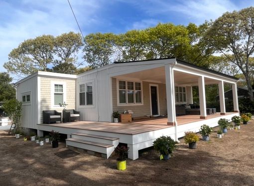 White tiny house with a large wooden deck and covered porch in a wooded yard
