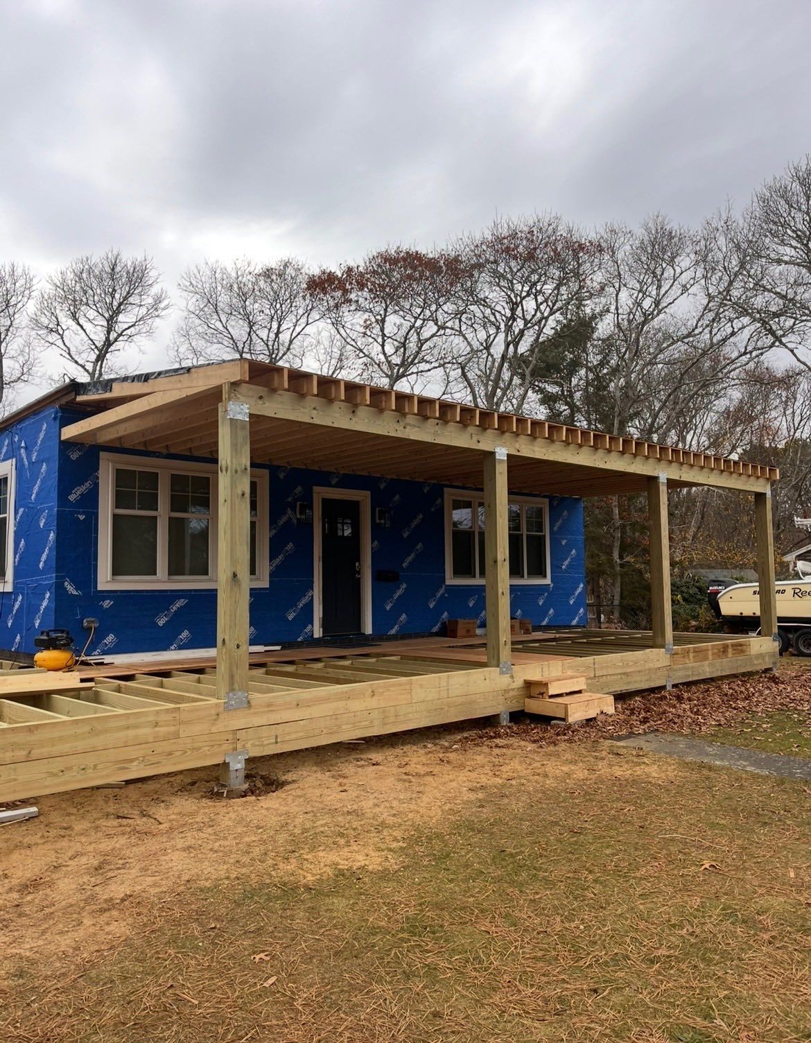 Blue house with a covered porch and wooden ramp, surrounded by leafless trees on a cloudy day