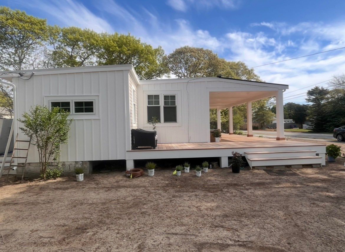 White mobile home with front porch and steps, set on a dirt lot under a blue sky.