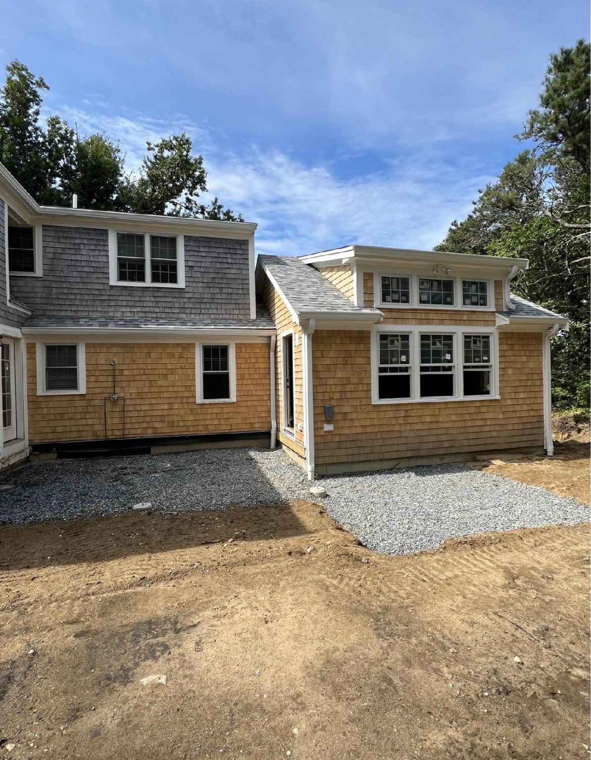 Two-story house exterior with tan siding, gray shingles, and gravel driveway under a blue sky