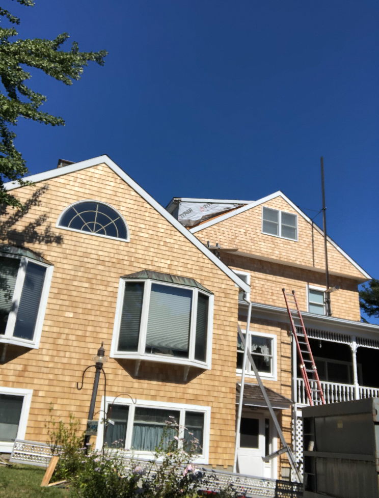 Tan-sided house with white-trimmed windows and a ladder against the upper porch under a clear blue sky