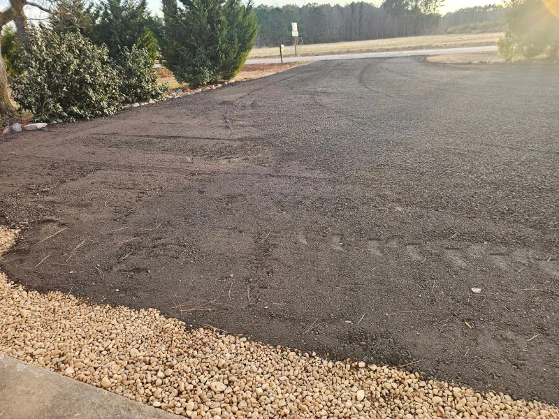 Gravel driveway with brown stones, adjacent to a gravel border and greenery, in an outdoor setting.