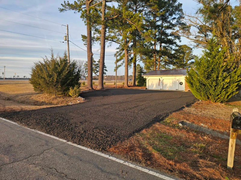 Driveway paved with dark asphalt leading to a white garage, trees in the background, beside a road.