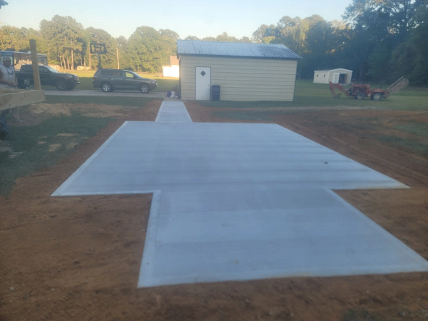 Newly poured concrete slab in a yard with a small building and vehicles in the background.