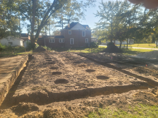 Construction site: foundation trenches and holes dug in dirt, house in background.