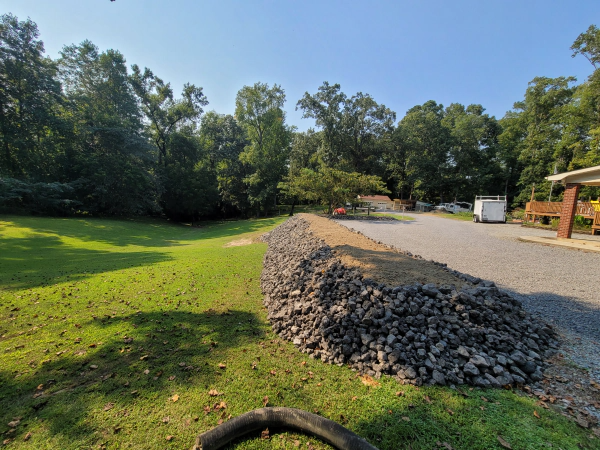 Grassy lawn with rock barrier next to gravel driveway; trees and a structure in the background.