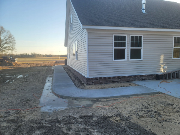 A concrete sidewalk curves around the side of a light gray house. Construction site.