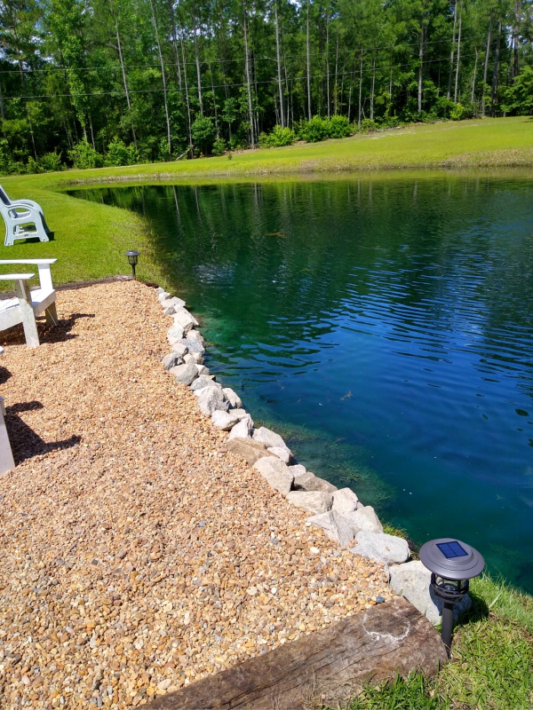 Pond with gravel edge, two white chairs, and trees in the background. The water is blue, and the sky is sunny.