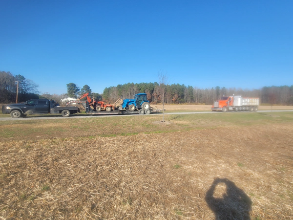 Truck towing tractor on flatbed trailer; passing semi-truck on road; sunny day.