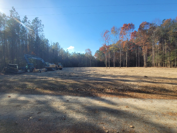 A cleared field bordered by trees; construction equipment sits in the distance on a sunny day.