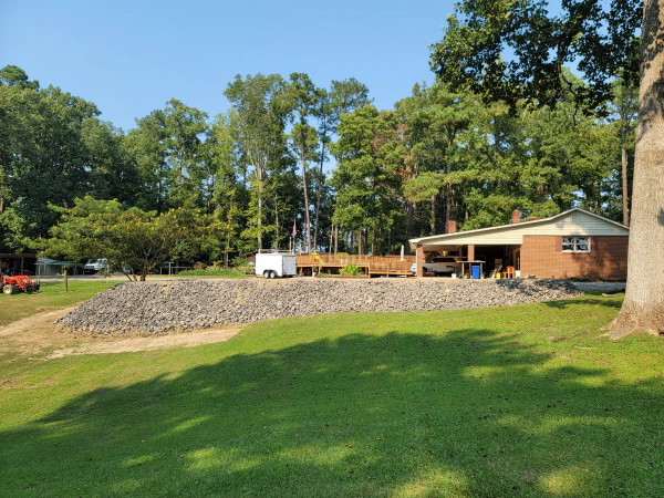 A grassy yard slopes up to a rocky berm with a house and trees in the background under a blue sky.