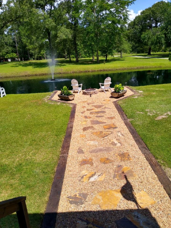 Stone pathway leading to a fire pit and seating by a pond with a fountain. Green lawn and trees.