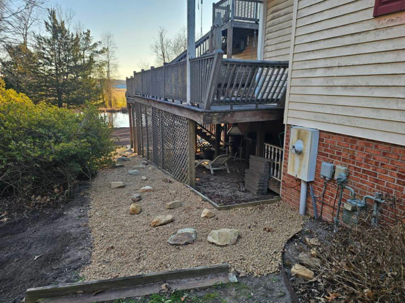 Side of a house with a deck. Gravel path with rocks leads to underneath deck. Electrical boxes on brick wall.