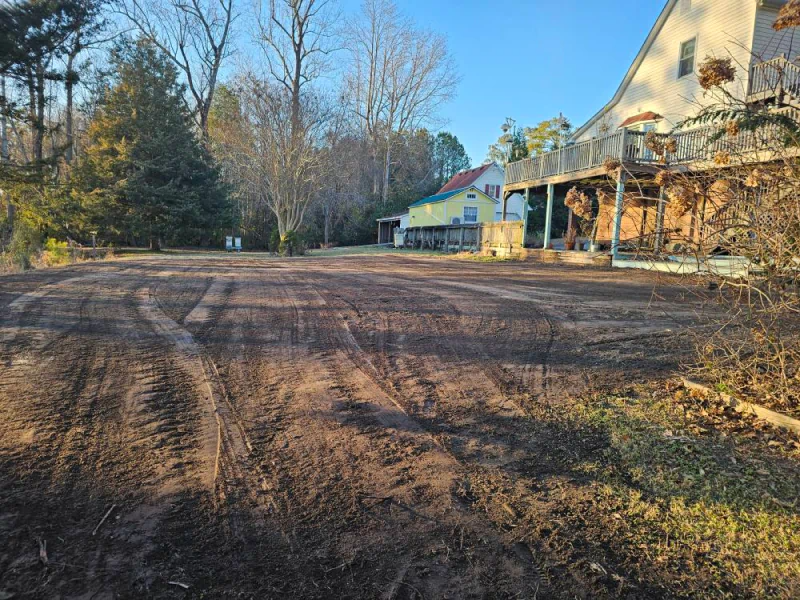 Dirt parking area in front of a two-story house with a deck and trees.