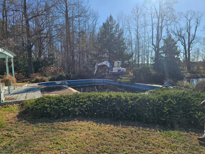 A small excavator is demolishing an old pool surrounded by overgrown bushes and trees on a sunny day.