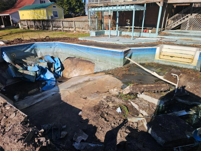 A dilapidated outdoor pool with damaged concrete and blue liner, in a yard.
