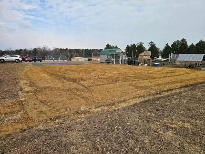 Large, cleared plot of land with brown earth, buildings, and parked cars under a cloudy sky.