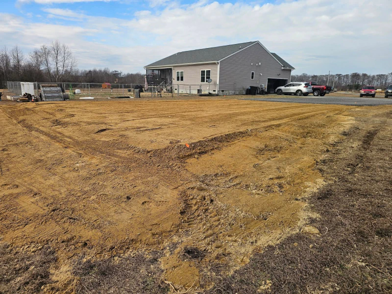 Brown dirt lot with tire tracks in front of a house with a gray exterior and a white truck parked in the driveway.
