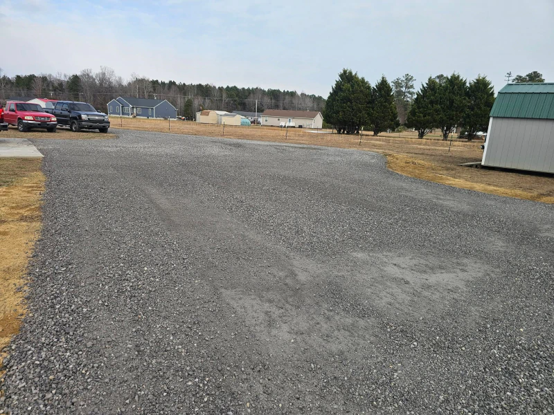 Gravel parking area with parked vehicles, shed, trees, and houses in the distance under a cloudy sky.