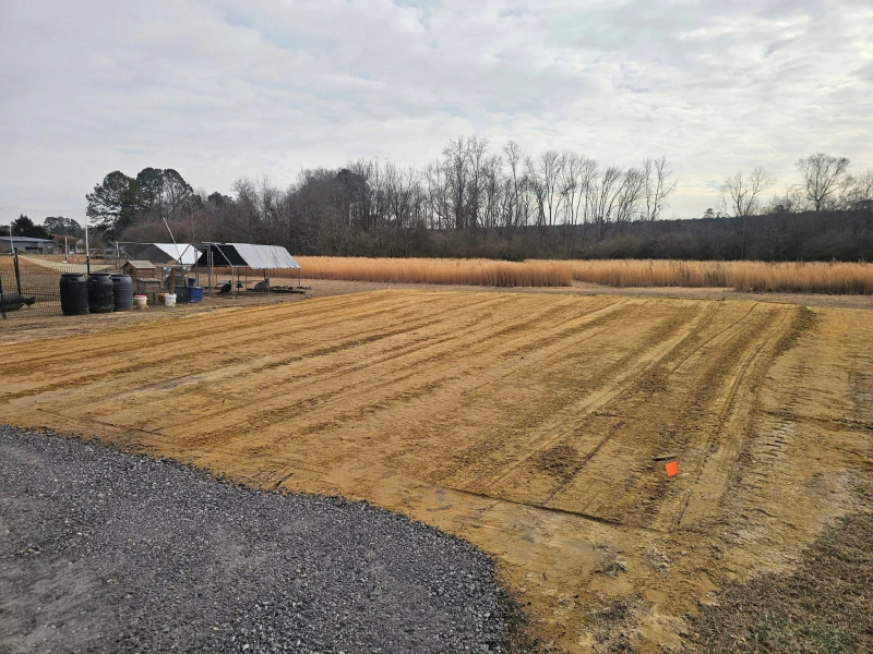 A plowed field, gravel path, barrels, and a small shelter under a cloudy sky.