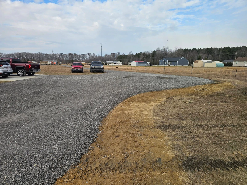 Gravel parking lot with vehicles and a few buildings in the background. The sky is cloudy.
