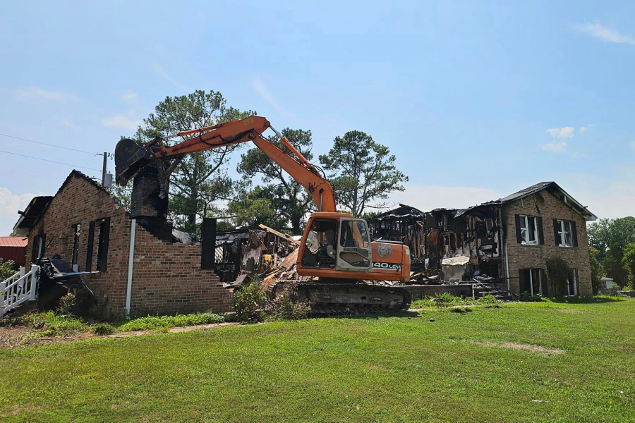 Excavator demolishing a burnt brick house on a green lawn under a blue sky.