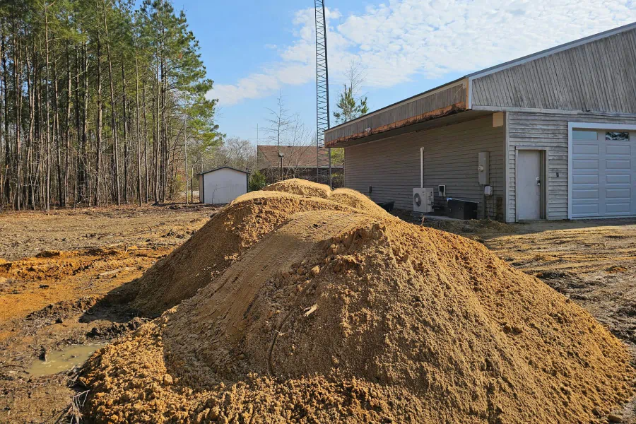 A large pile of sand on a dirt lot in front of a building and a forest of trees.