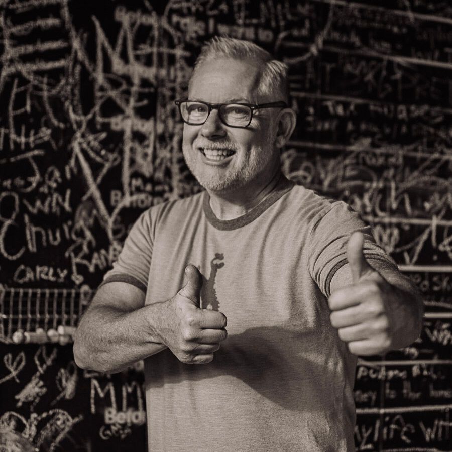 Smiling man with glasses giving two thumbs up in front of a chalkboard wall