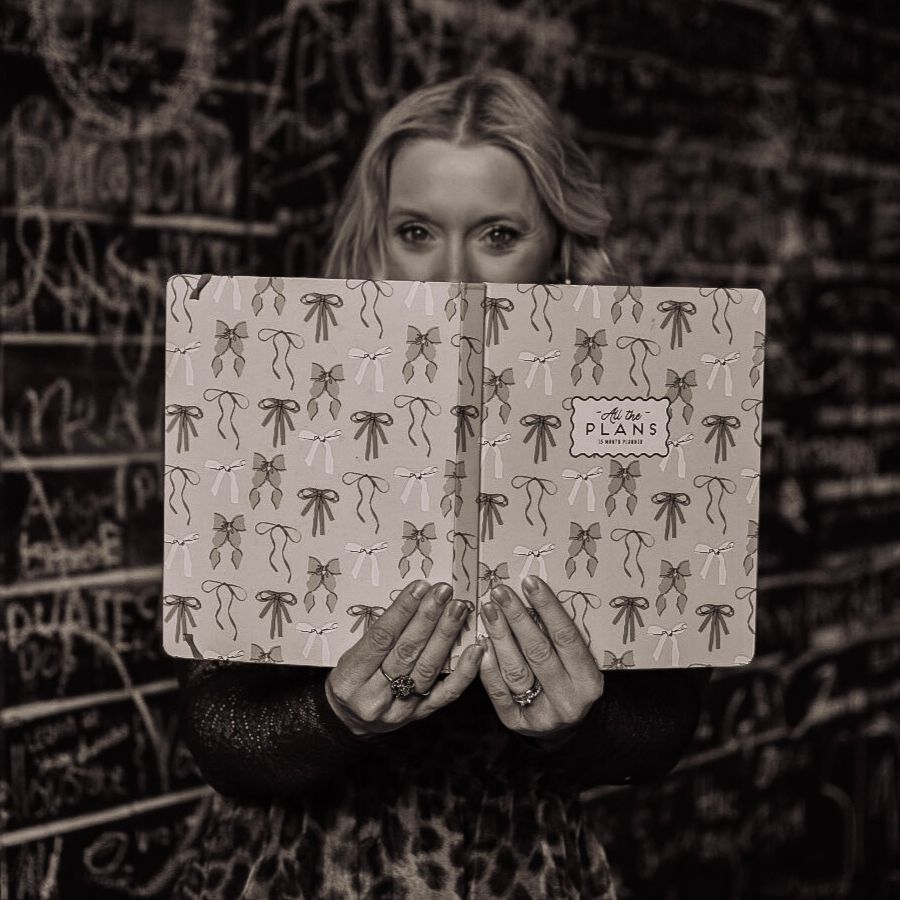 Woman holding a patterned notebook over her face, with only her eyes visible against a chalkboard wall.