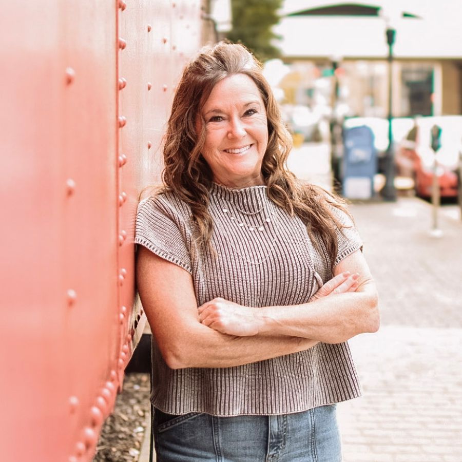 Smiling woman leaning against a red wall on a city street, arms crossed.