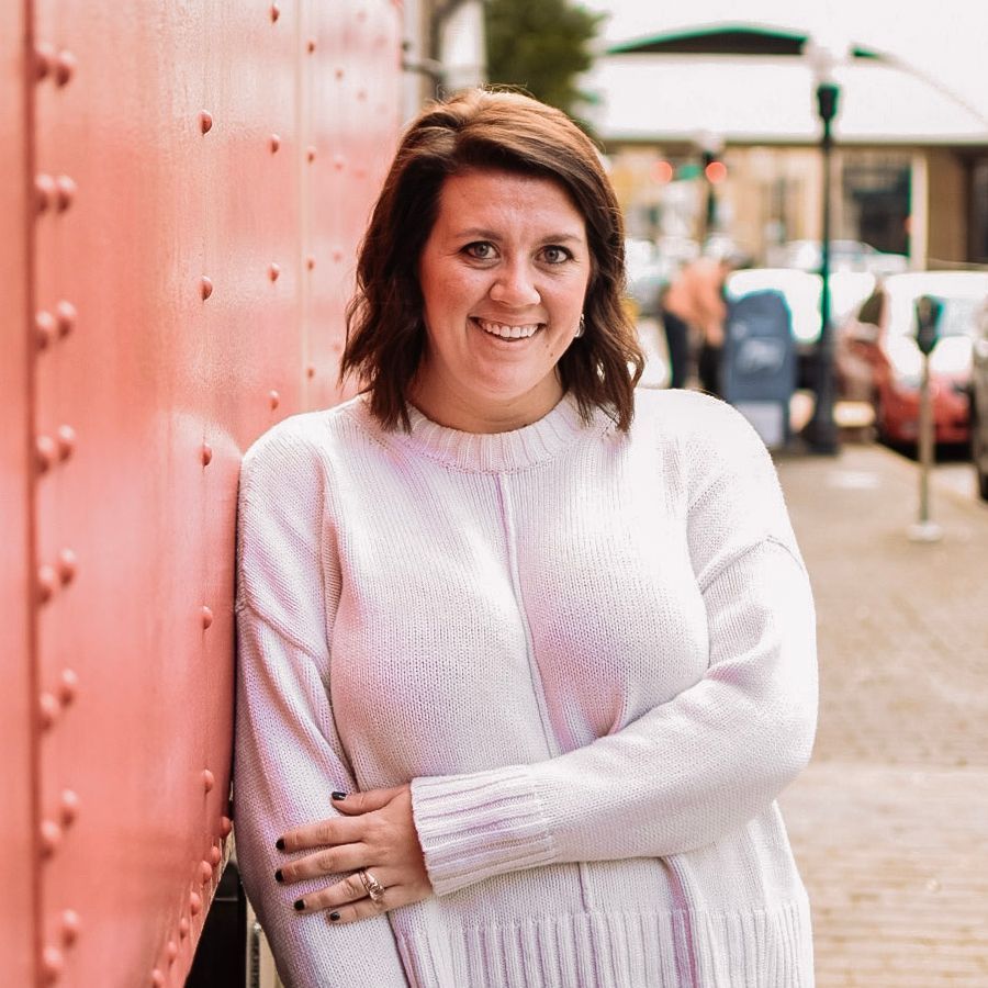 Woman in a white sweater leaning against a red wall on a city sidewalk, smiling at the camera