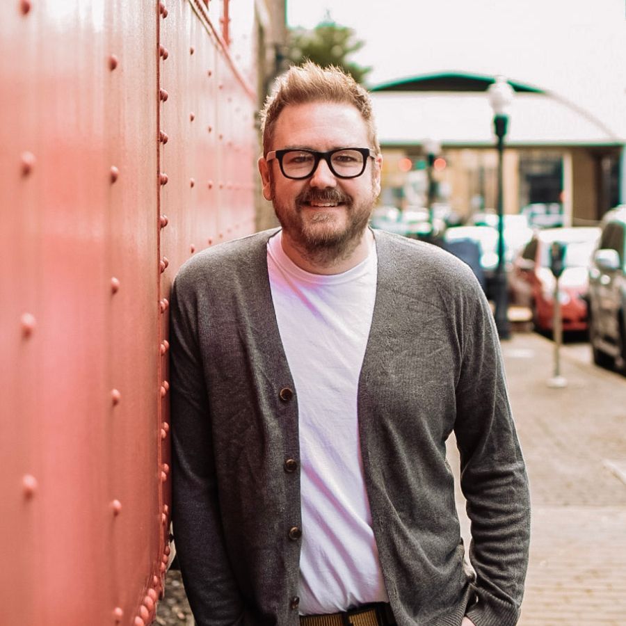 Man leaning against a red wall on a city sidewalk, wearing glasses, cardigan, and white T-shirt.