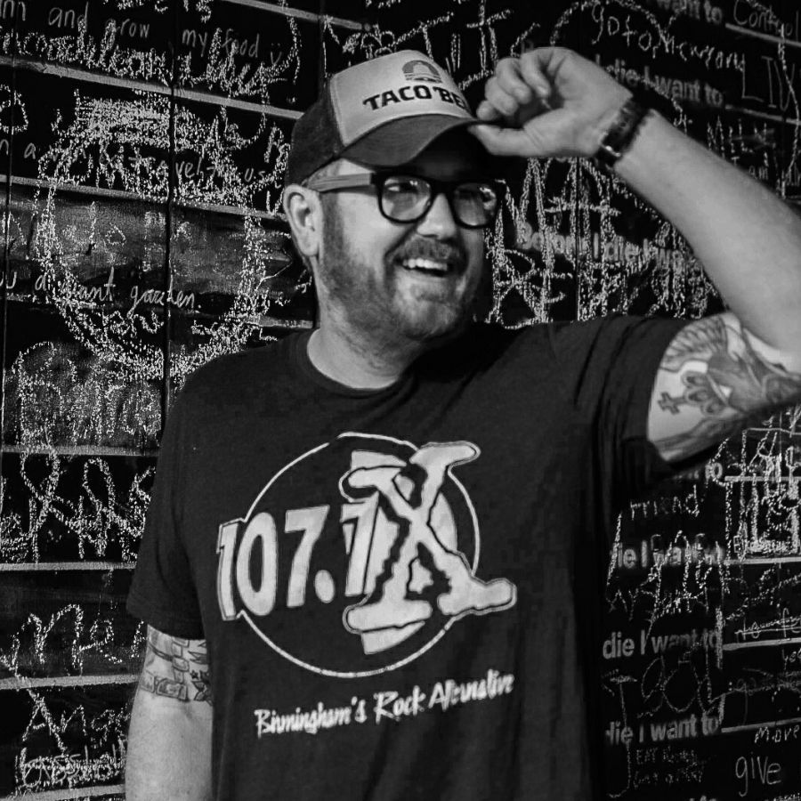 Smiling man in glasses and cap adjusting brim, wearing 107.7 X T-shirt in front of a chalkboard wall